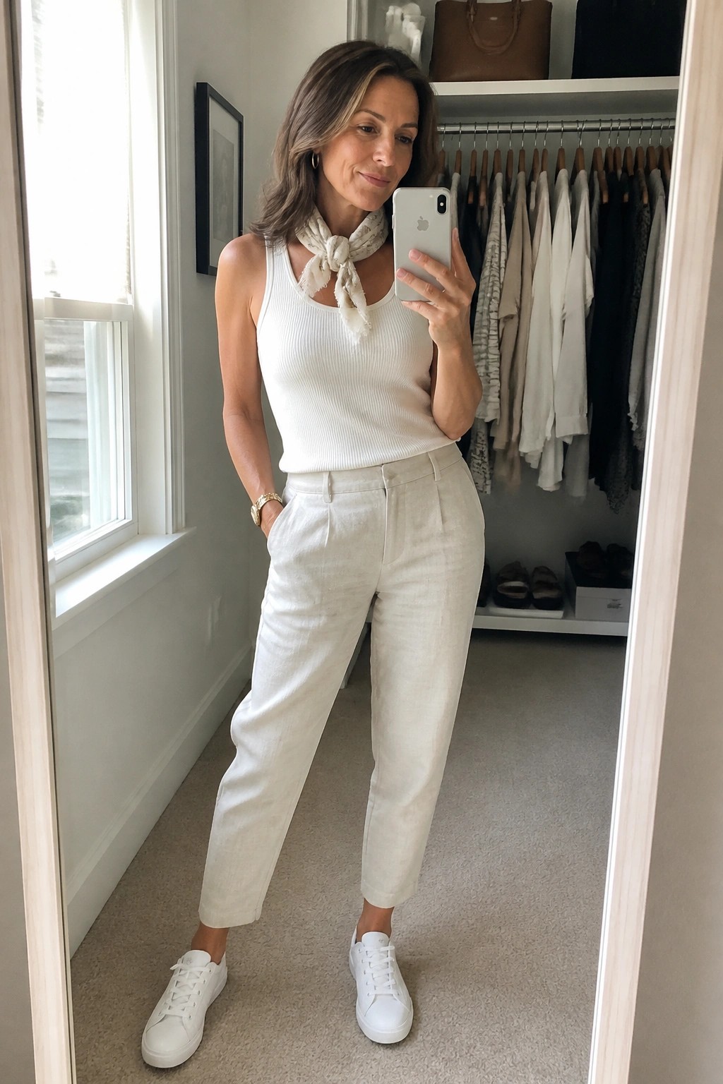 Woman over 50 wearing an all-white outfit with a ribbed tank top, linen straight-leg pants, white sneakers, and a light scarf tied at the neck while taking a mirror selfie in a closet.