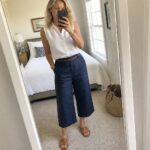 Woman wearing a white sleeveless button front shirt tucked into navy wide leg cropped pants with a brown belt and matching slide sandals while taking a mirror selfie.