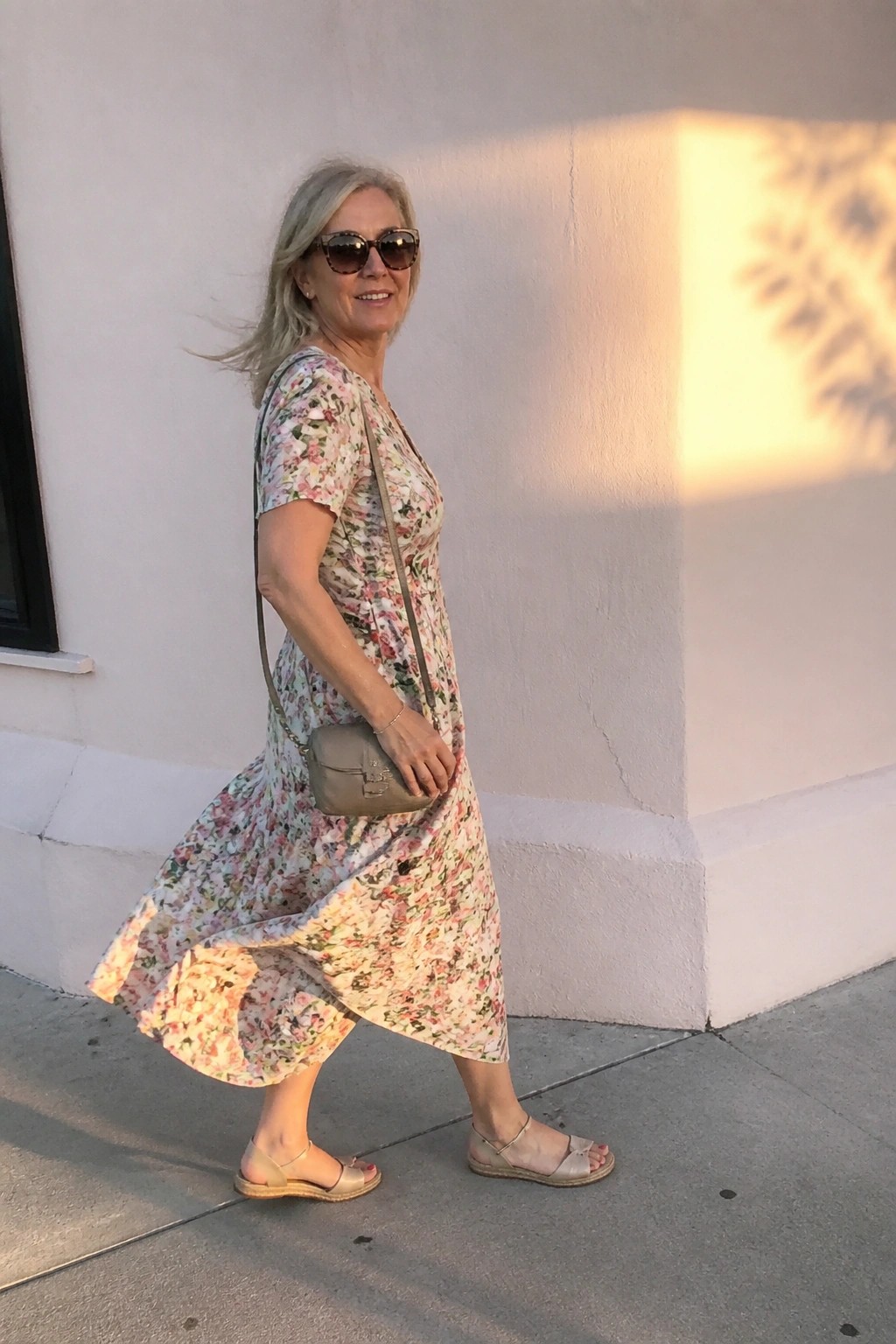 Woman in floral midi dress with crossbody bag walking by white wall in sunlight.