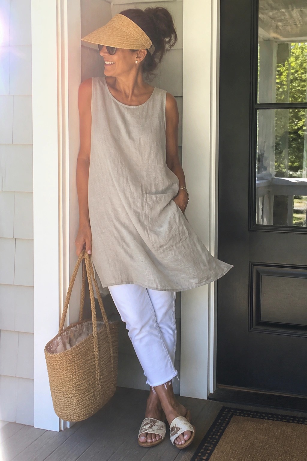 Woman over 50 wearing a sleeveless light beige linen tunic with front pockets over white cropped jeans, paired with a straw visor, woven tote bag, and floral slide sandals.