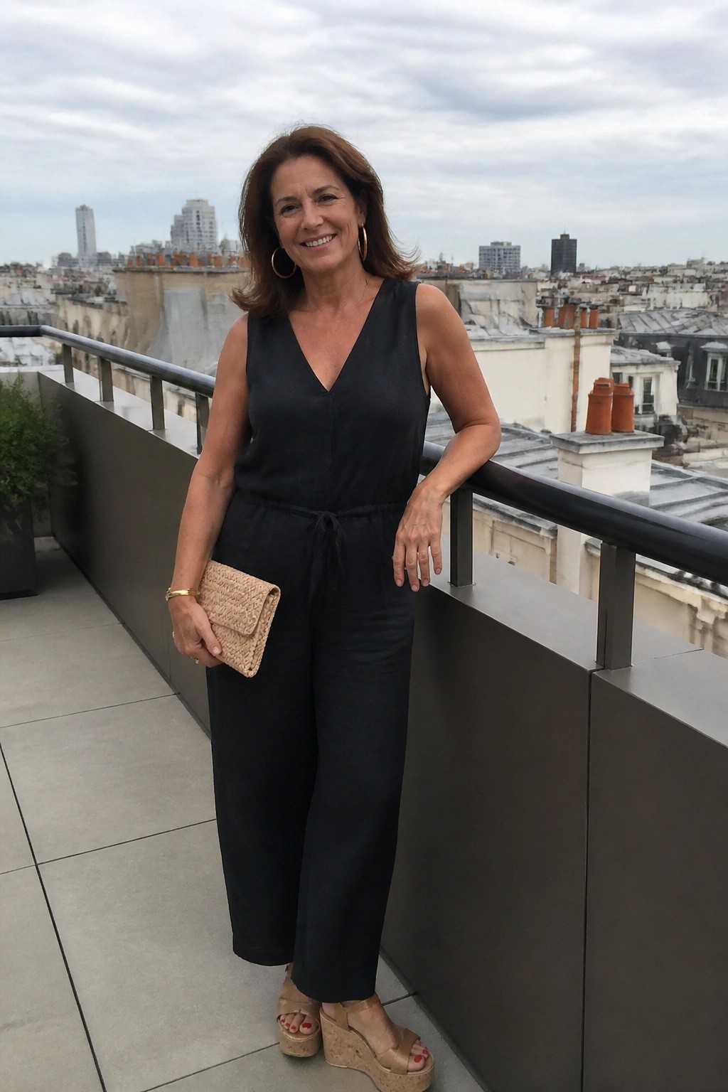 Woman in black jumpsuit smiling on balcony with city skyline, holding woven clutch.