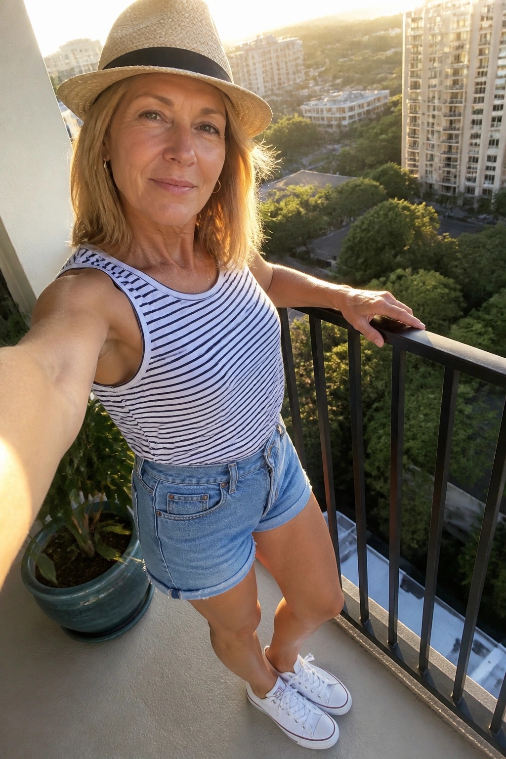 Woman over 50 in a navy striped sleeveless tank top tucked into light blue denim shorts with white sneakers and a straw hat.