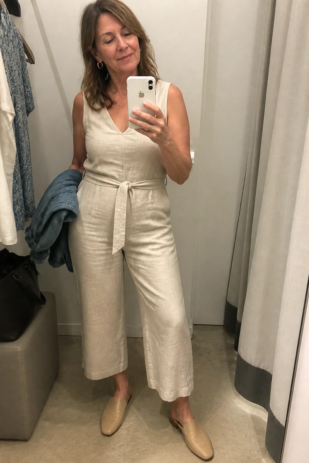 Woman in beige linen jumpsuit taking mirror selfie in fitting room holding denim jacket