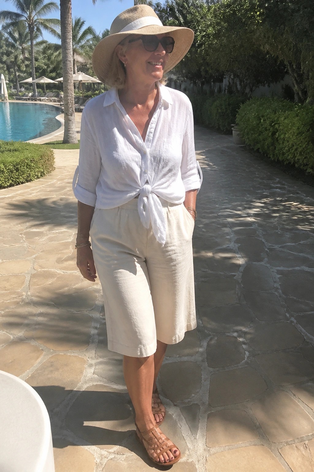 Woman in tied white shirt and beige shorts with straw hat near pool
