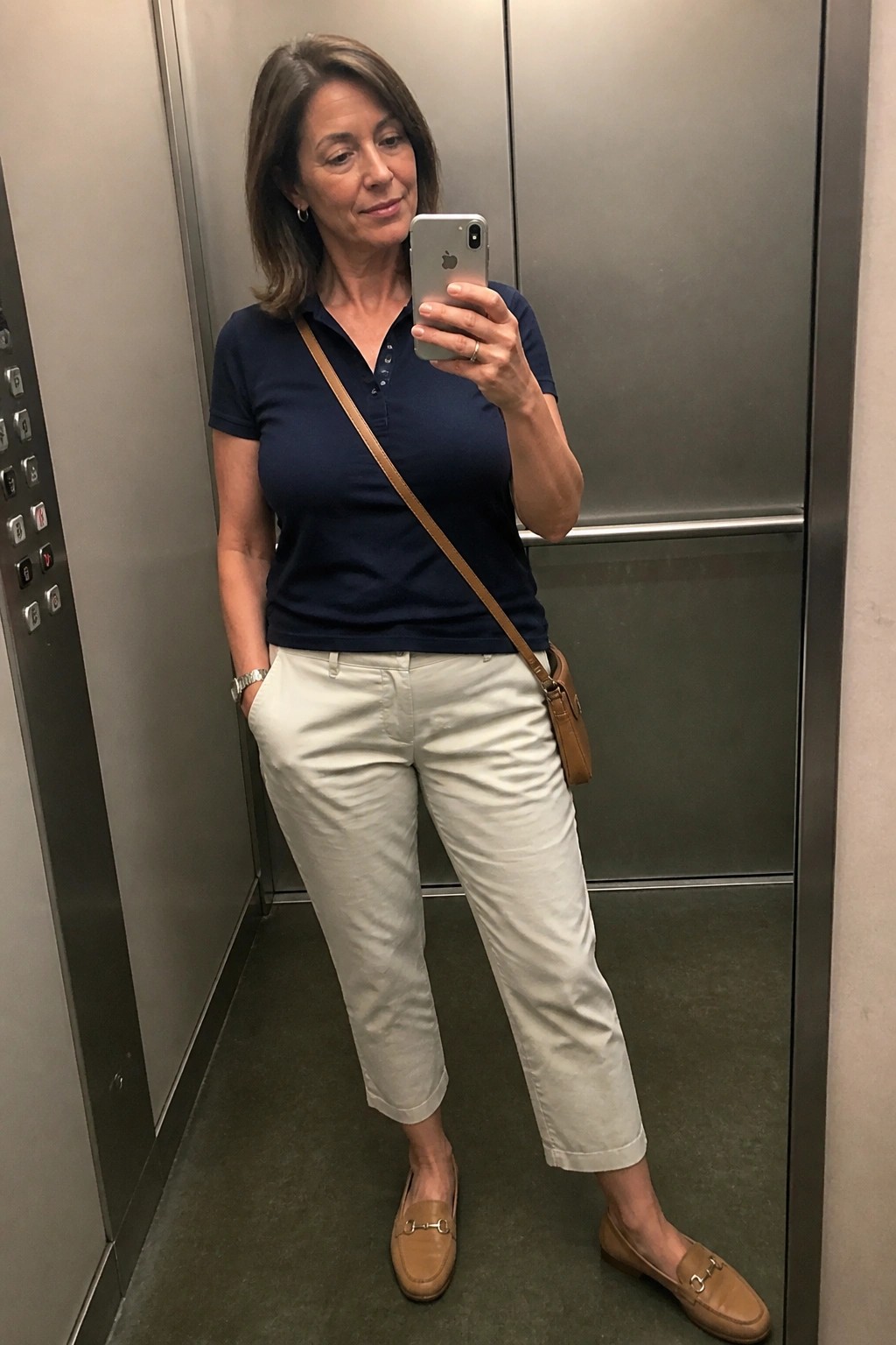 Woman over 50 in navy polo shirt tucked into cropped beige pants with tan loafers and brown crossbody bag taking elevator mirror selfie