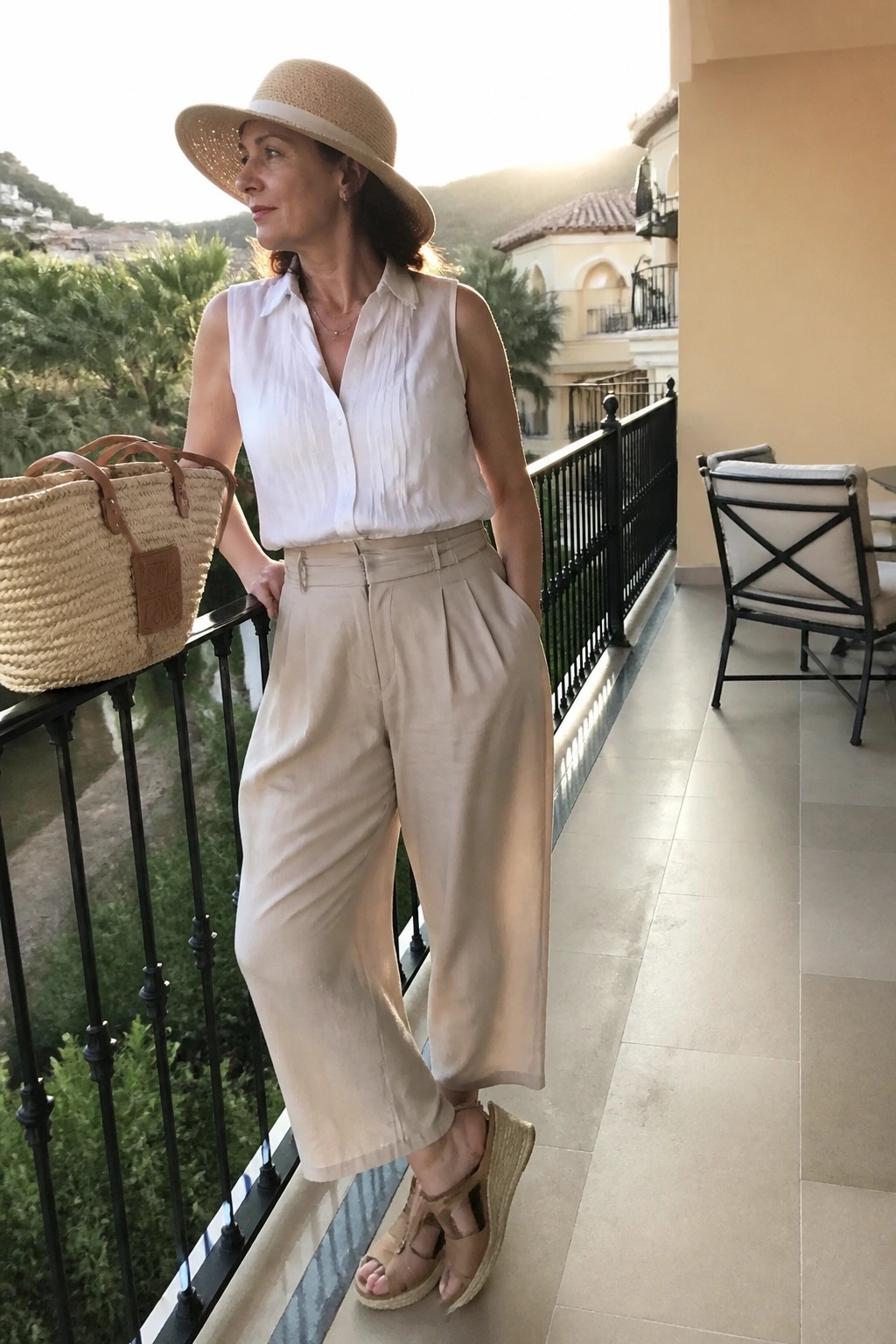 Woman in white sleeveless shirt and beige trousers with straw hat on balcony