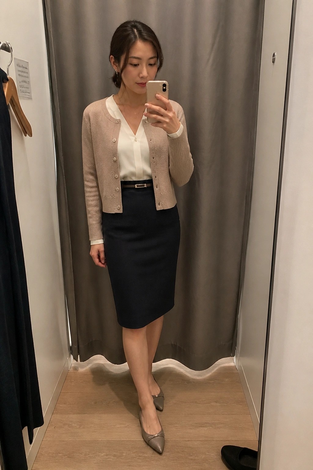 Woman in beige cardigan and navy skirt taking mirror selfie in fitting room