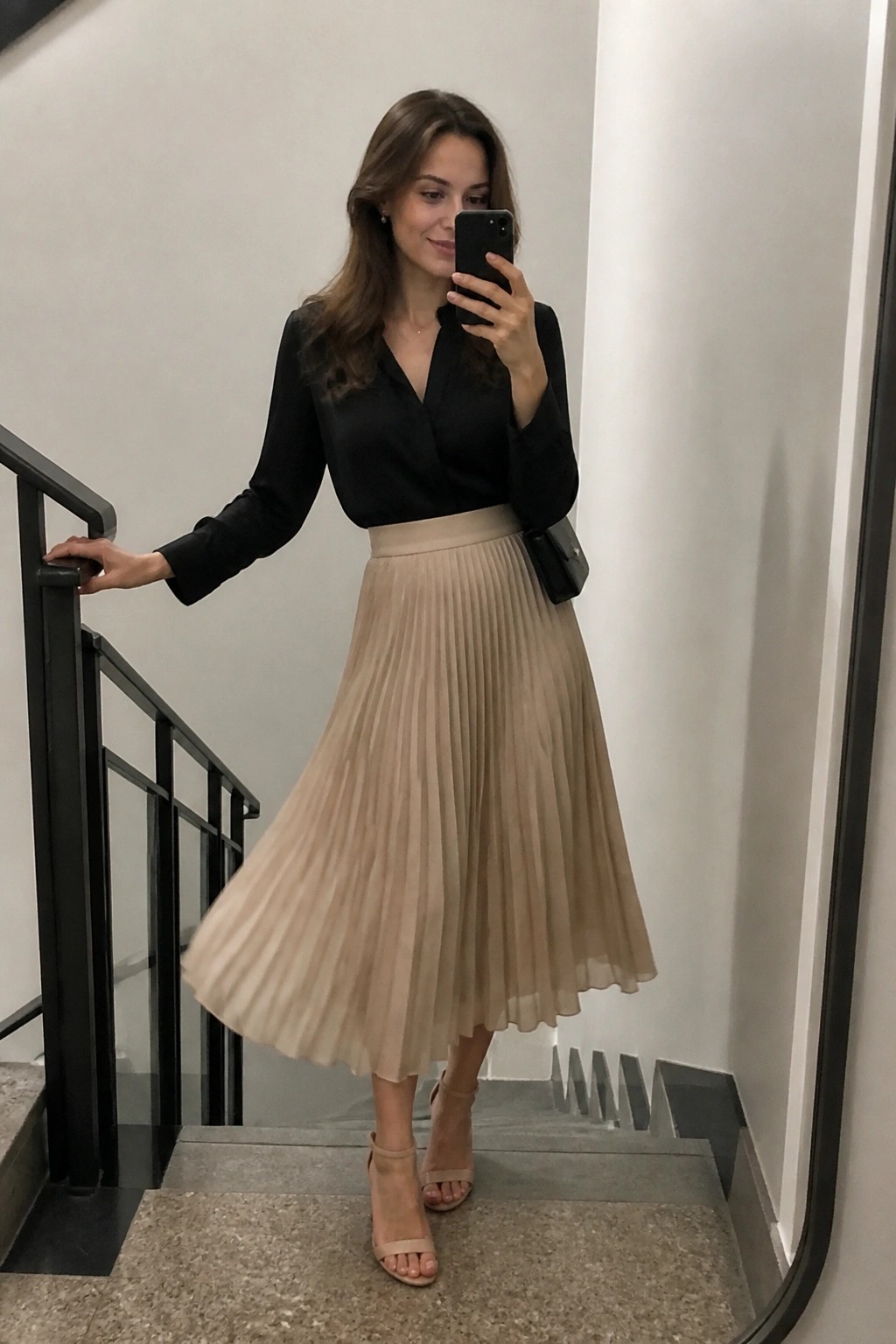 Woman wearing a black wrap blouse tucked into a beige pleated midi skirt with nude block heel sandals and a small black bag while taking a mirror selfie on stairs.