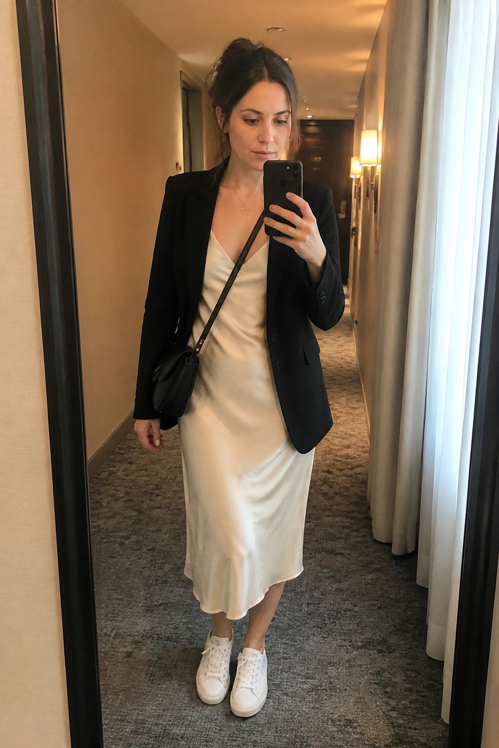 Woman in black blazer over cream satin slip dress paired with white sneakers and black crossbody bag taking mirror selfie in hallway.