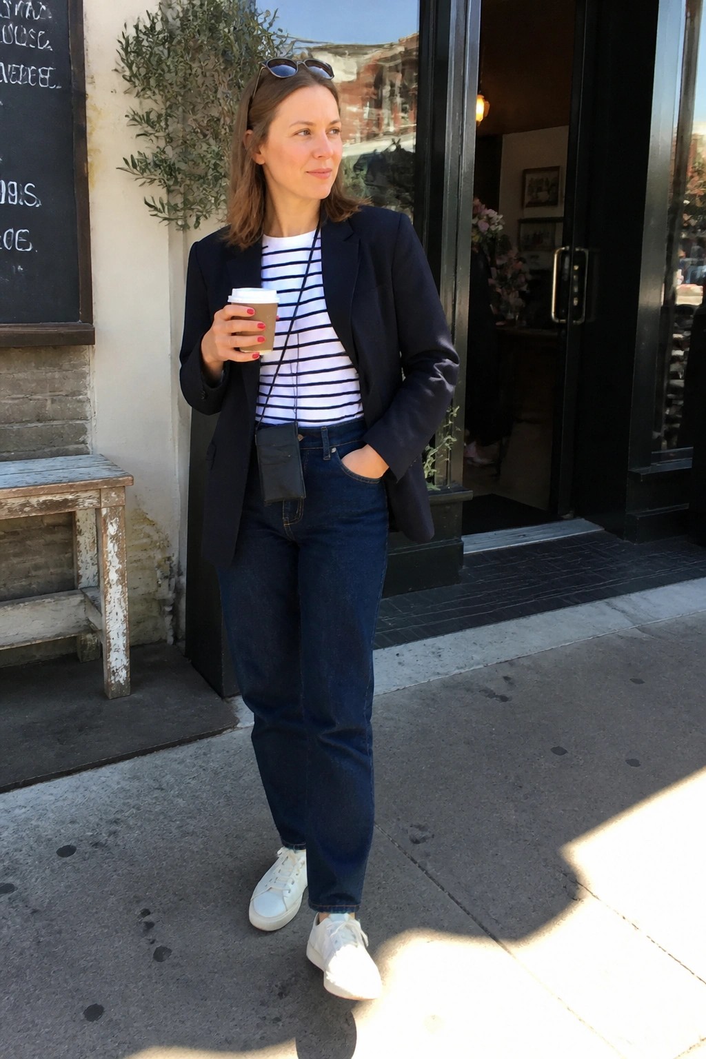Woman in navy blazer and striped shirt holds coffee outside shop entrance.