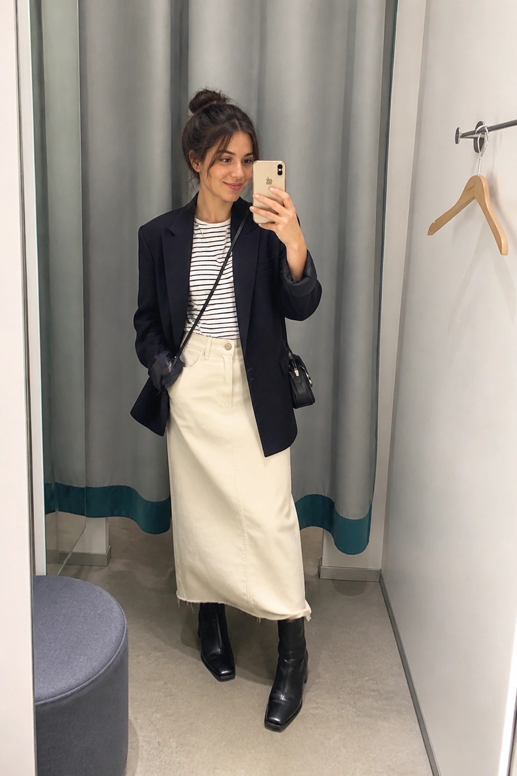 Woman in navy blazer, striped top, and cream denim skirt taking mirror selfie in fitting room