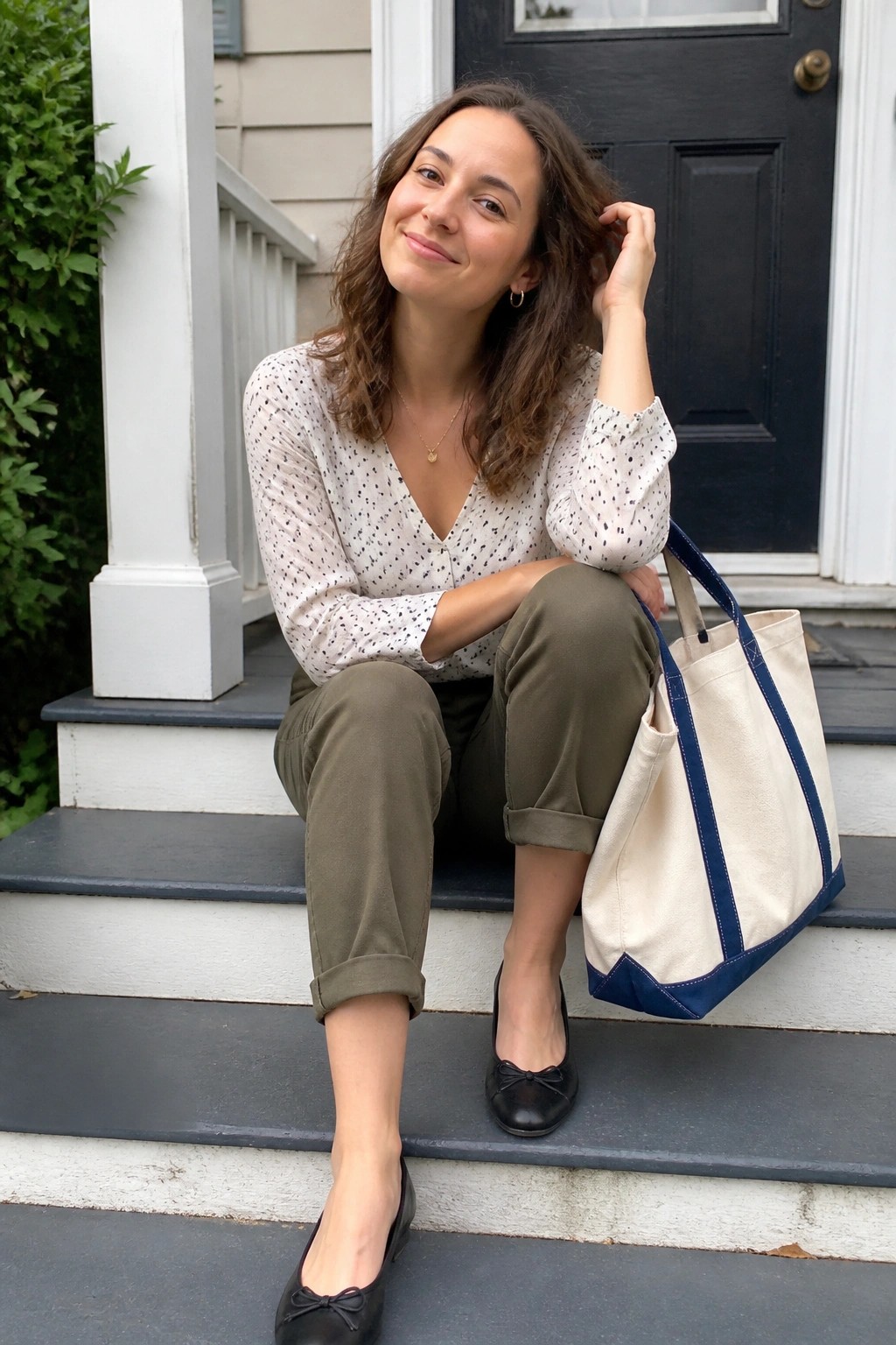 Woman seated on steps wearing a white long sleeve polka dot blouse tucked into olive green cuffed pants black bow ballet flats and carrying a cream canvas tote with blue trim.