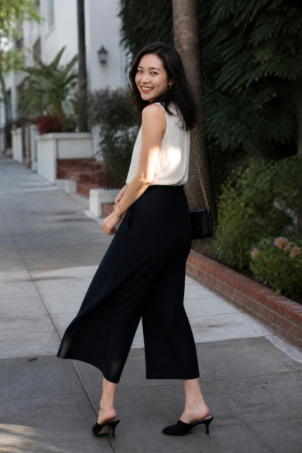 Woman smiling over shoulder in white top and black wide-leg pants on sidewalk