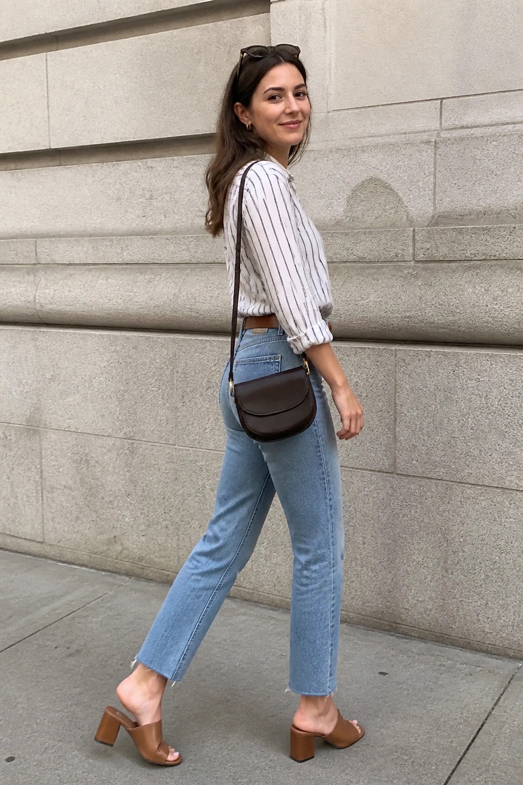 A woman in a white and black striped button-up shirt tucked into light blue straight-leg jeans with frayed hems, wearing brown heeled mules and carrying a brown leather crossbody bag.