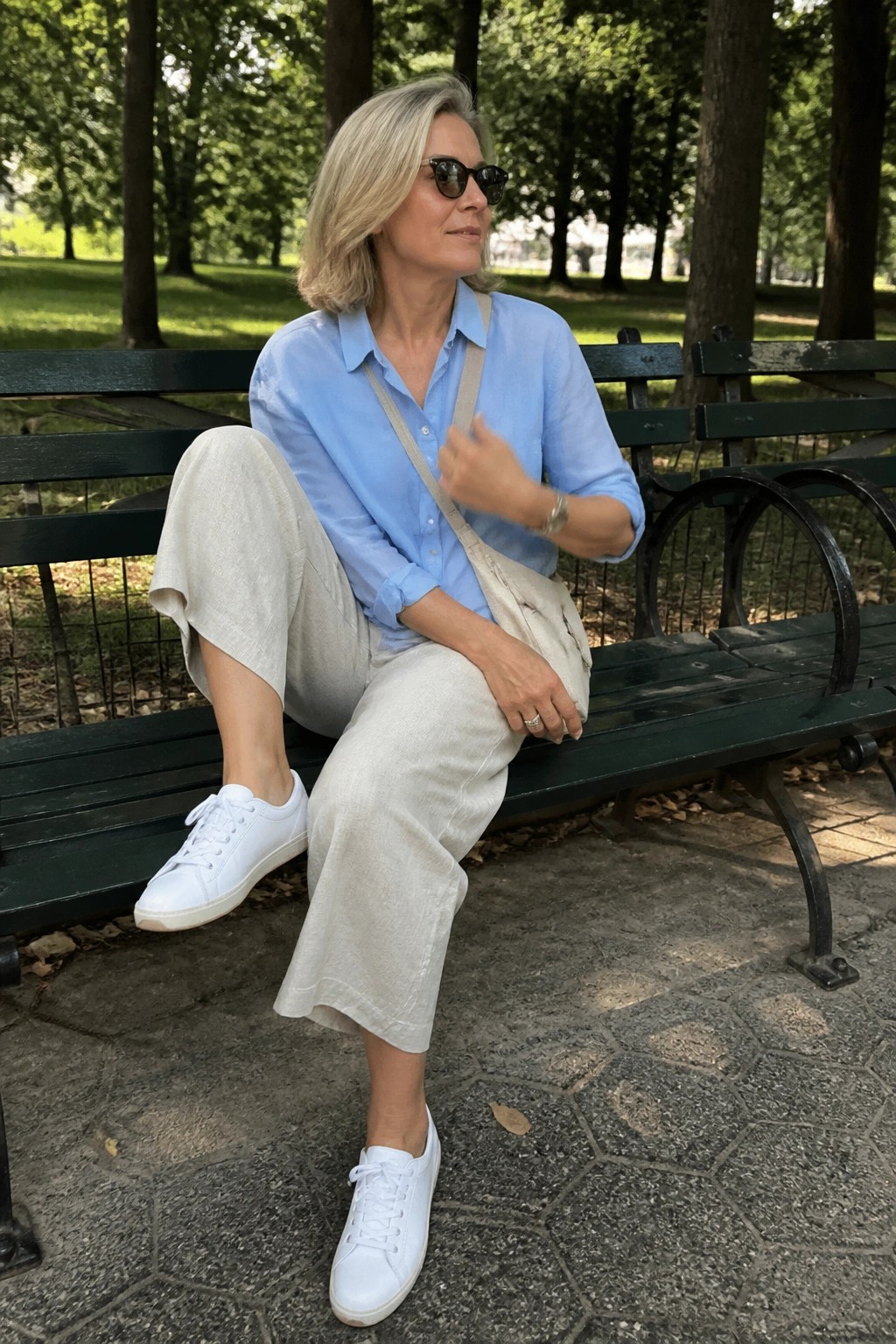 Woman over 50 wearing light blue button down shirt tucked into wide beige pants with white sneakers and beige crossbody bag while seated on a park bench.