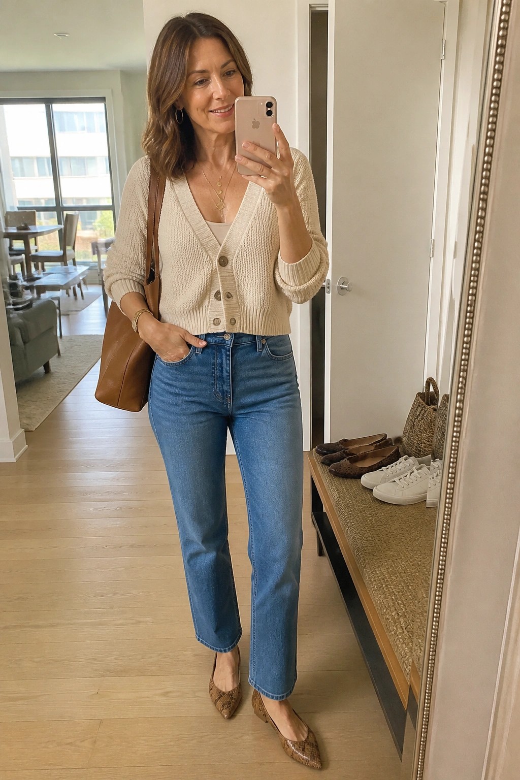 Woman in beige cardigan and blue jeans taking mirror selfie with brown tote bag
