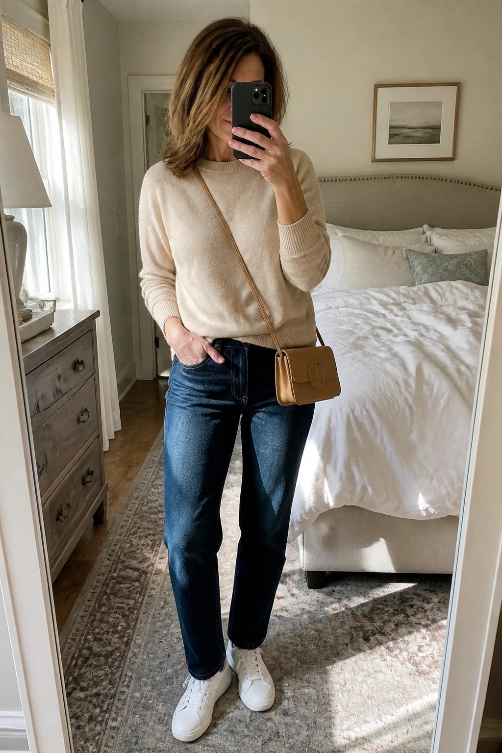 Woman in beige sweater and jeans taking mirror selfie in bedroom with tan bag