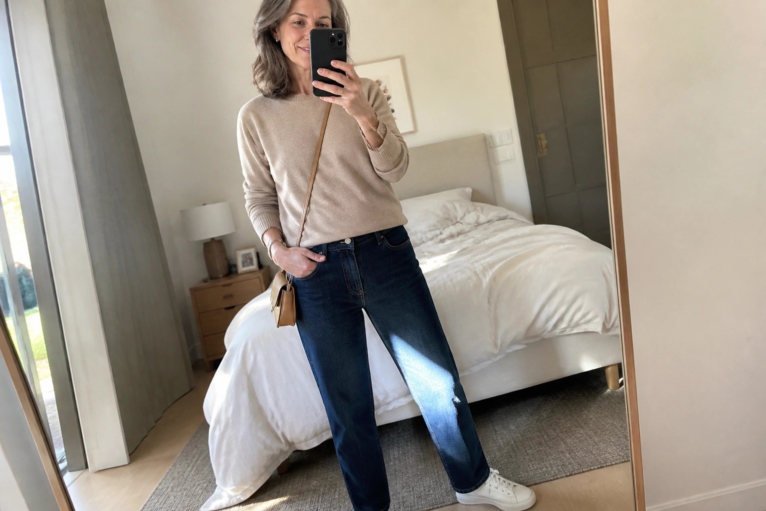 Woman in beige sweater and jeans taking mirror selfie in bedroom with tan bag
