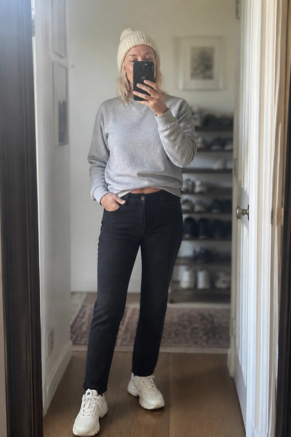 Woman in gray sweatshirt and black jeans taking mirror selfie indoors