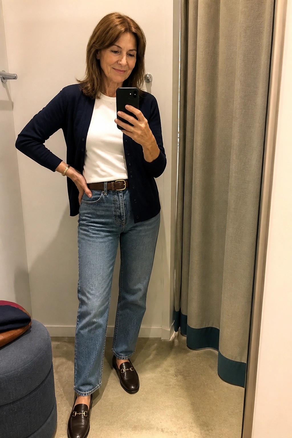 Woman over 50 wearing a navy cardigan over a white t-shirt with blue straight-leg jeans, brown belt, and brown loafers while taking a mirror selfie in a fitting room.