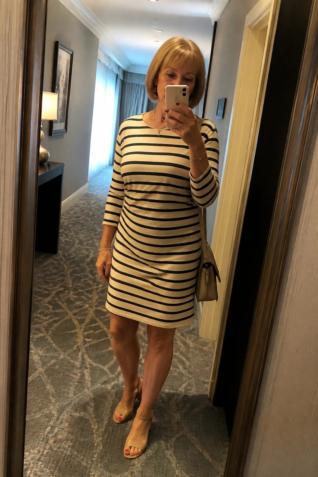 Woman in striped dress taking mirror selfie in hallway with beige purse