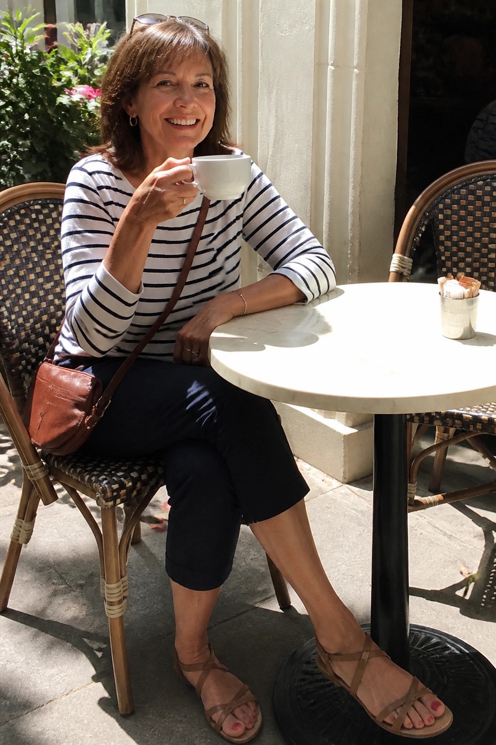 Woman wearing navy and white striped top with navy capri pants, brown leather crossbody bag, and brown strappy sandals while seated at a cafe table holding a coffee cup
