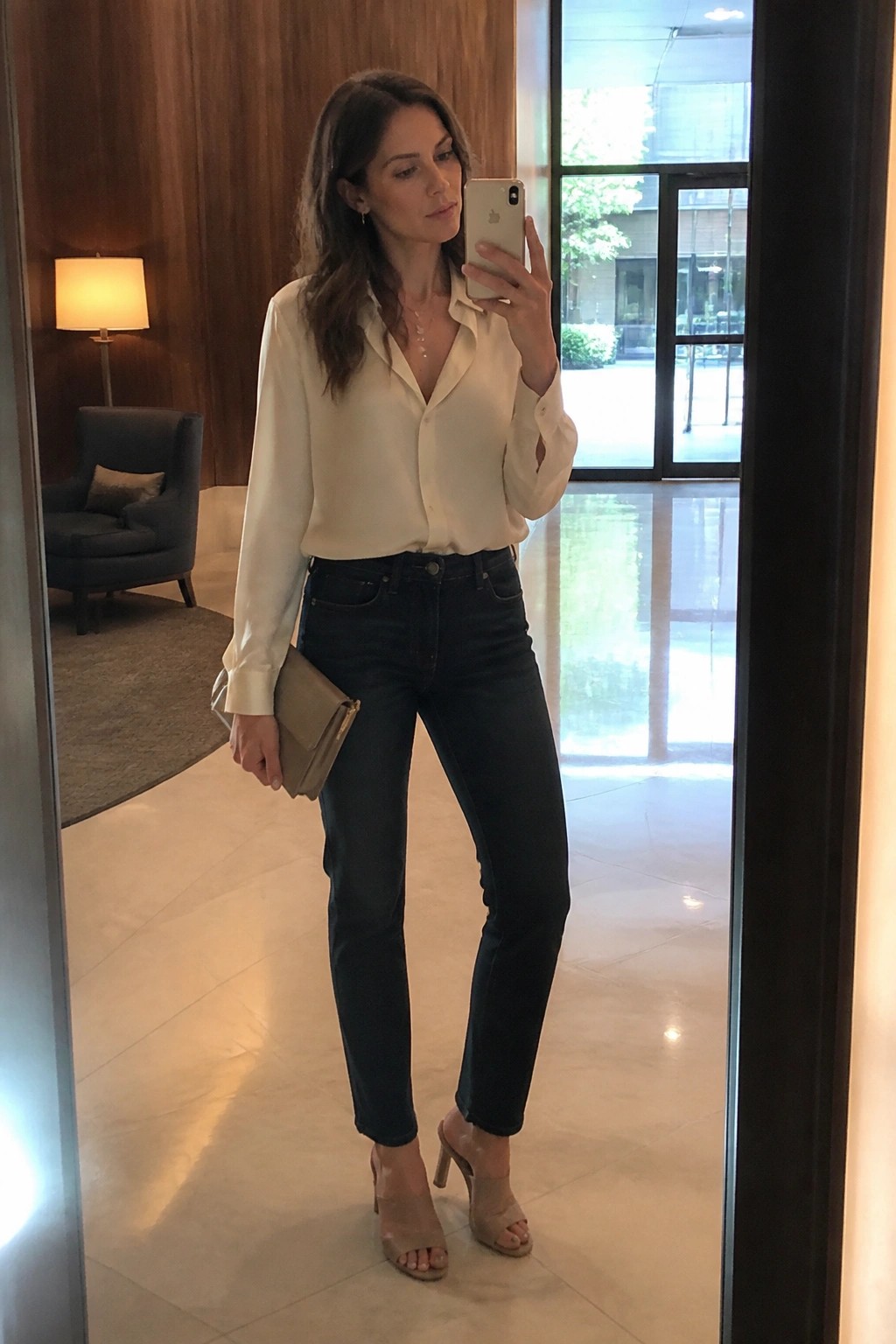 Woman in cream blouse and dark jeans taking mirror selfie with clutch and phone