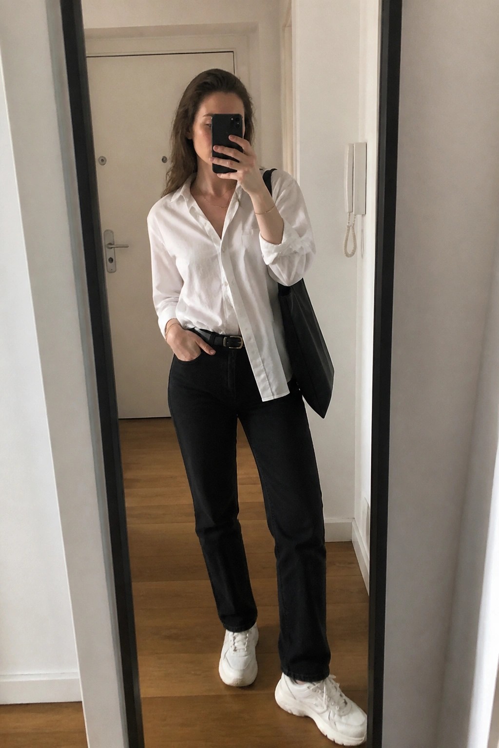 Woman in white button-down shirt and black jeans taking mirror selfie with phone and bag