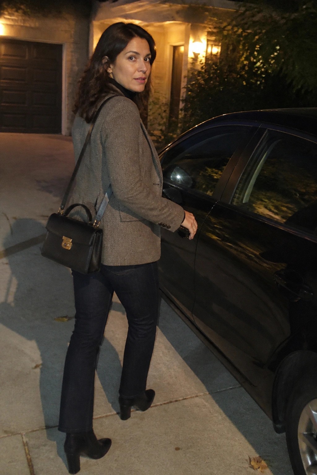 Woman in gray blazer and jeans with black handbag beside car at night.