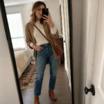 Woman in cream sweater and tan cardigan taking bedroom mirror selfie with phone.