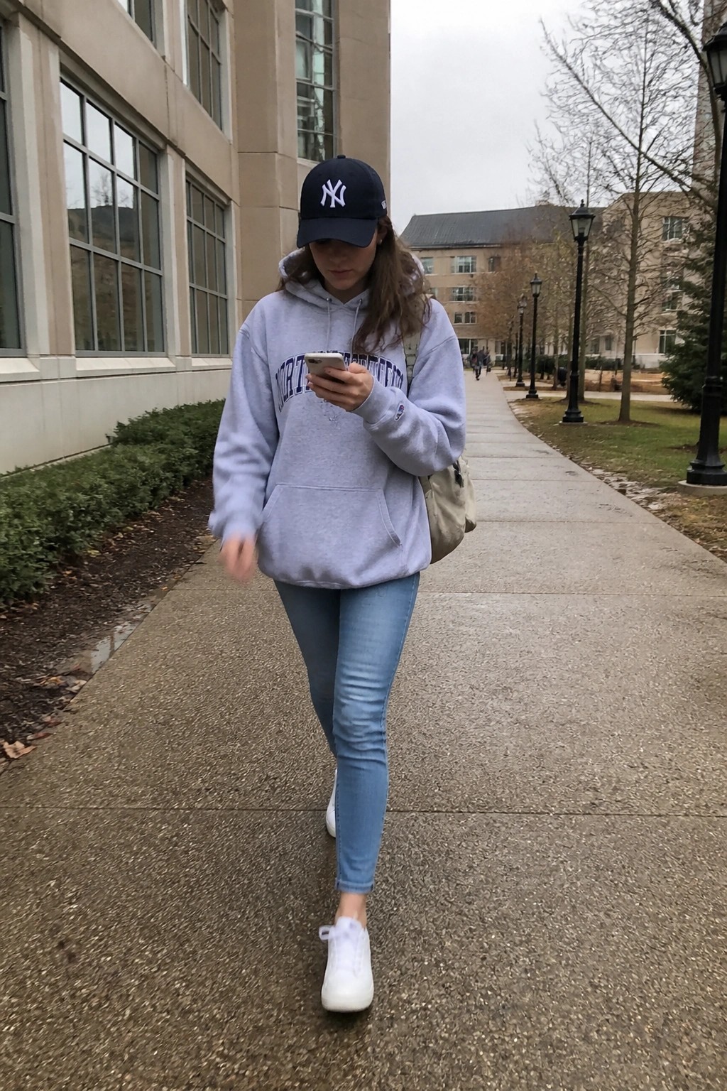 Woman in gray hoodie and Yankees cap checking phone on campus walkway.