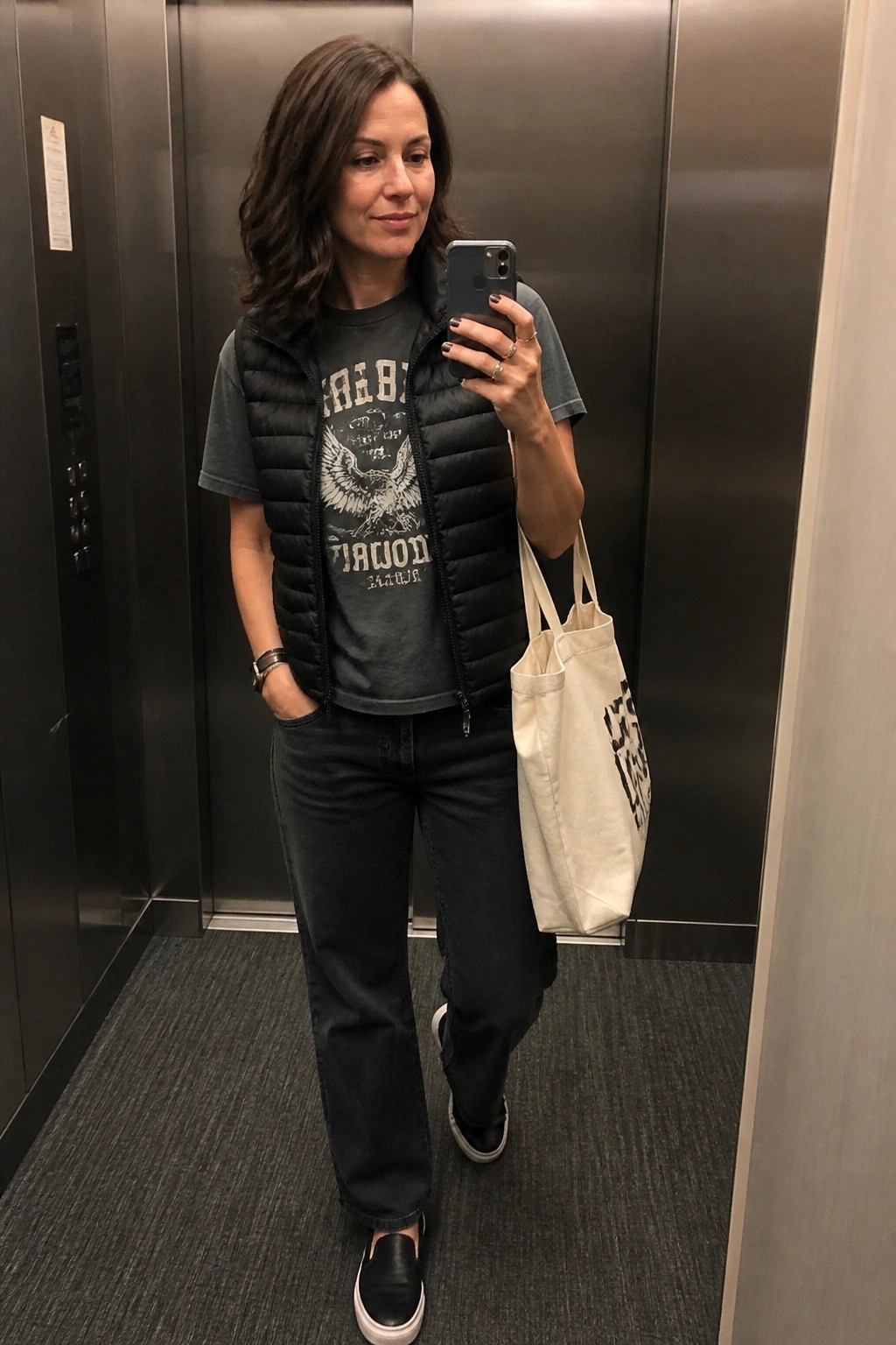 Woman in black puffer vest taking selfie with tote bag in elevator