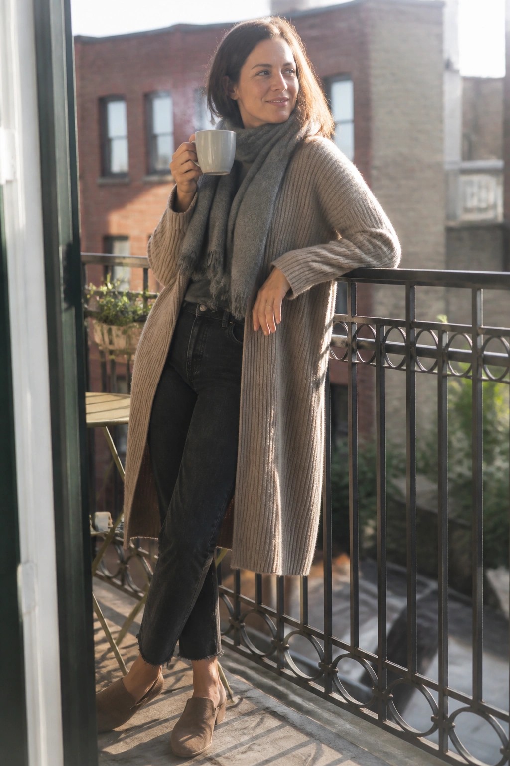 Woman in beige cardigan and gray scarf holding steaming mug on balcony.