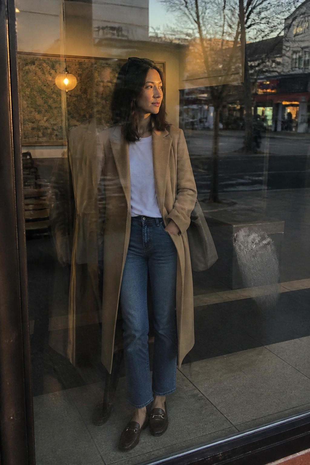 Woman in beige coat and jeans standing by storefront window at dusk.