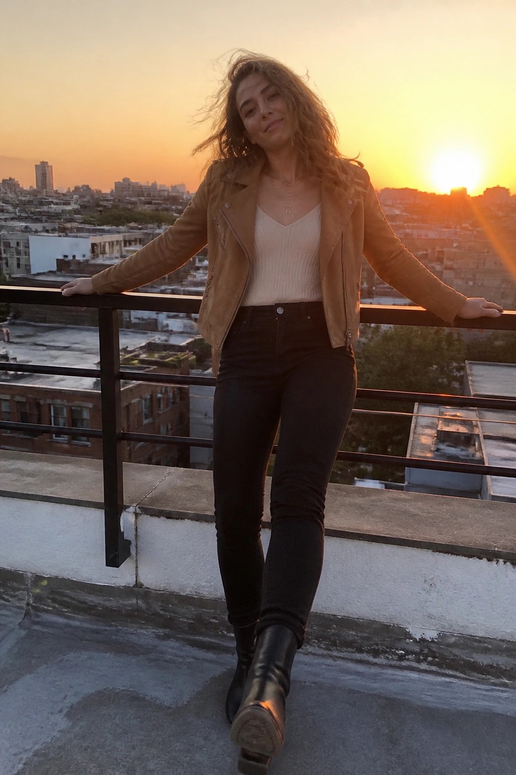 Woman in brown jacket and black jeans posing on rooftop at sunset