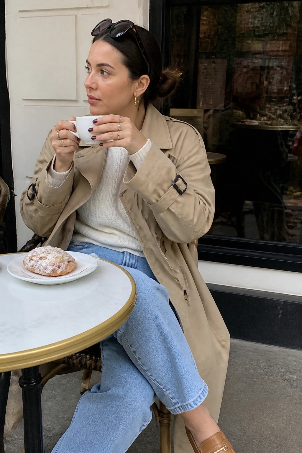 Woman seated outdoors wearing an open beige trench coat over a white cable knit sweater and light blue cropped jeans with brown loafers, holding a white coffee cup, with dark nail polish and gold rings visible.