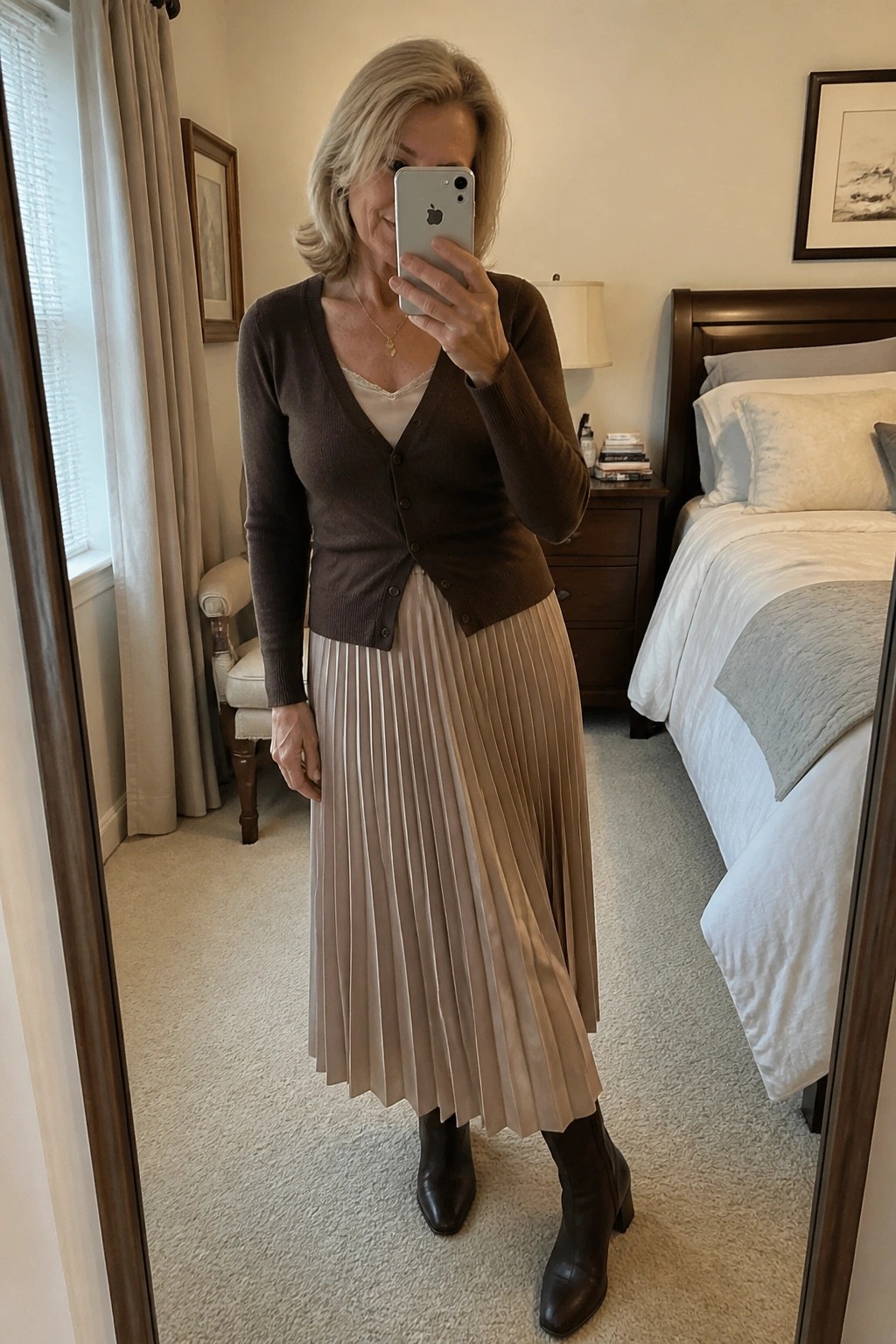 Woman in brown cardigan and pleated beige skirt taking bedroom mirror selfie
