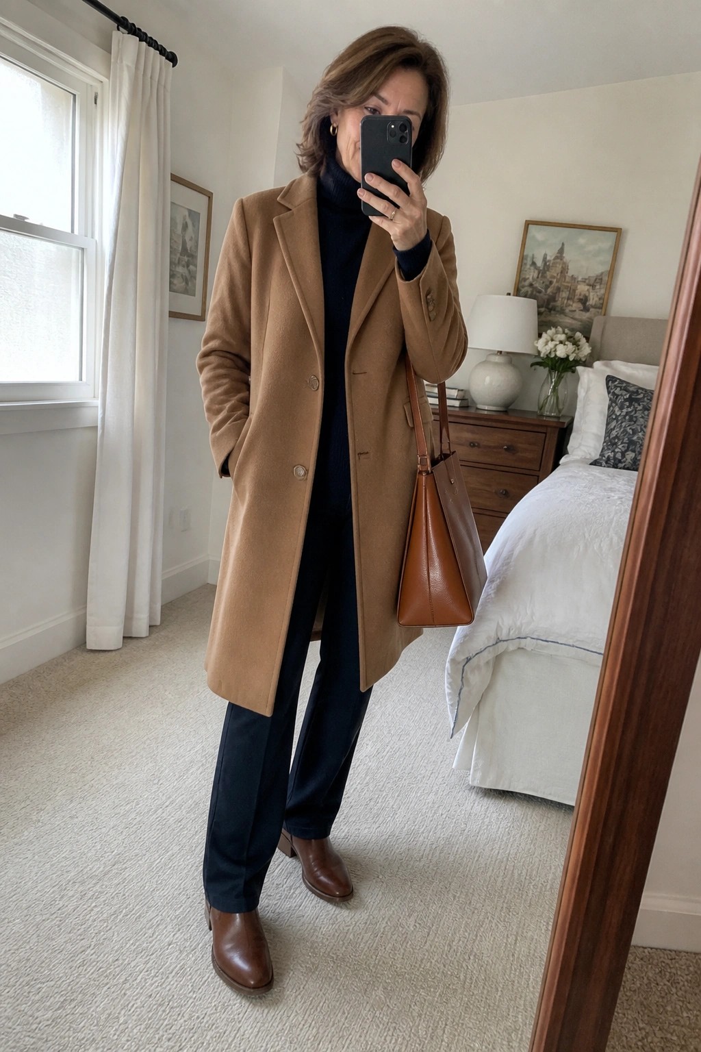 Woman over 60 wearing a long camel coat buttoned over a navy turtleneck and matching navy pants, paired with brown leather ankle boots and a matching brown tote bag, holding a phone for a mirror selfie in a bedroom setting.