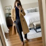 Woman over 60 wearing a long camel coat buttoned over a navy turtleneck and matching navy pants, paired with brown leather ankle boots and a matching brown tote bag, holding a phone for a mirror selfie in a bedroom setting.