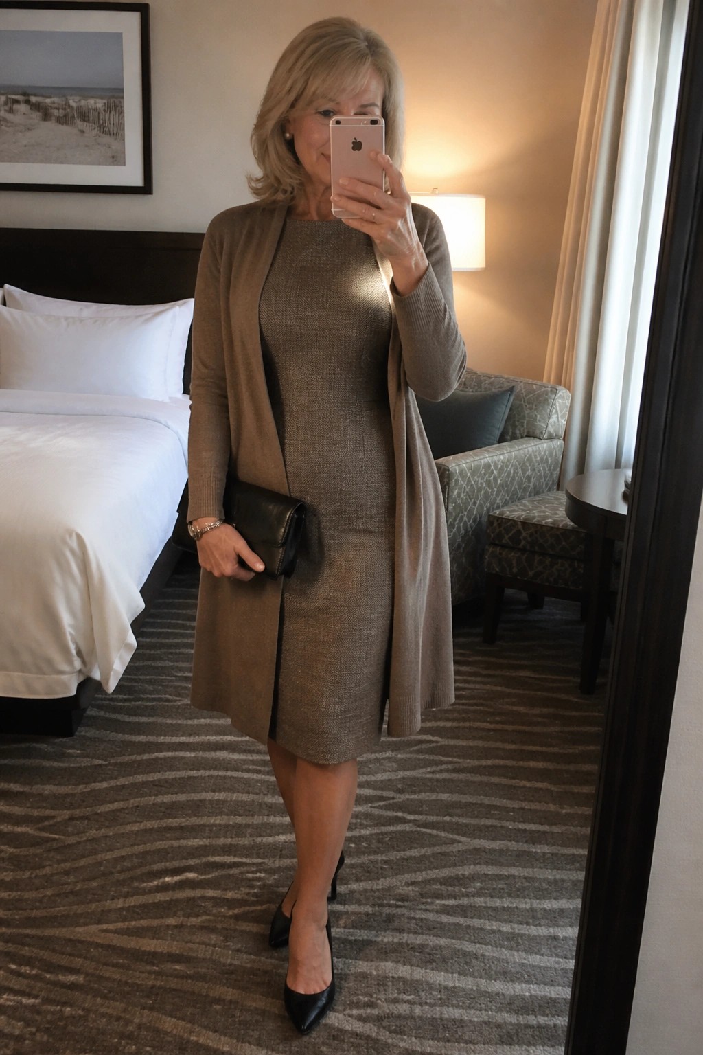 Woman in taupe cardigan over sparkly dress taking mirror selfie in hotel room