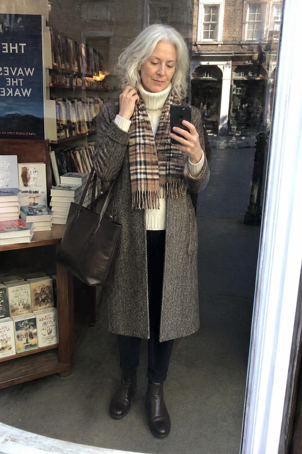 White-haired woman in herringbone coat and plaid scarf takes selfie outside bookstore.