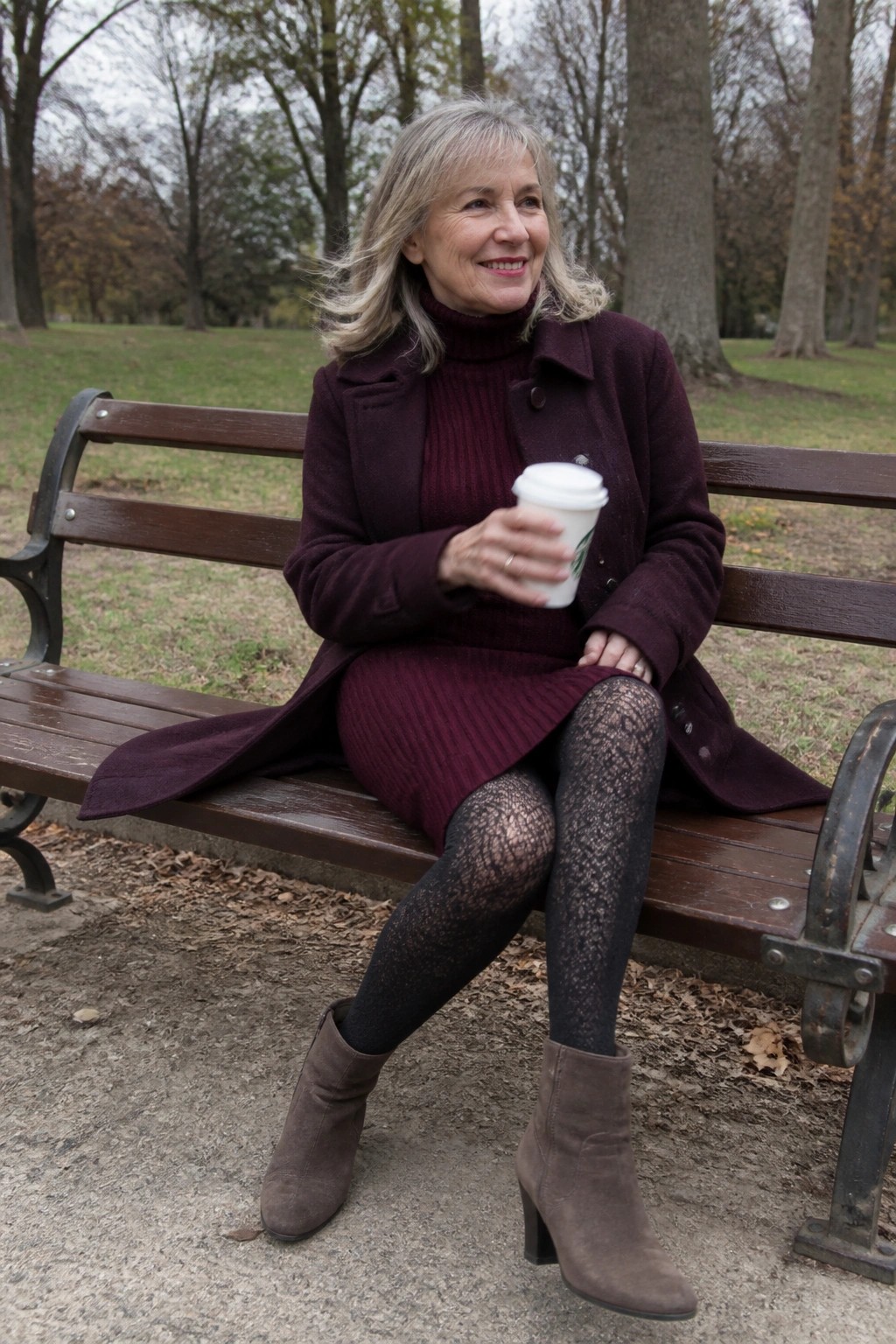 Smiling woman in maroon coat holding Starbucks cup on park bench