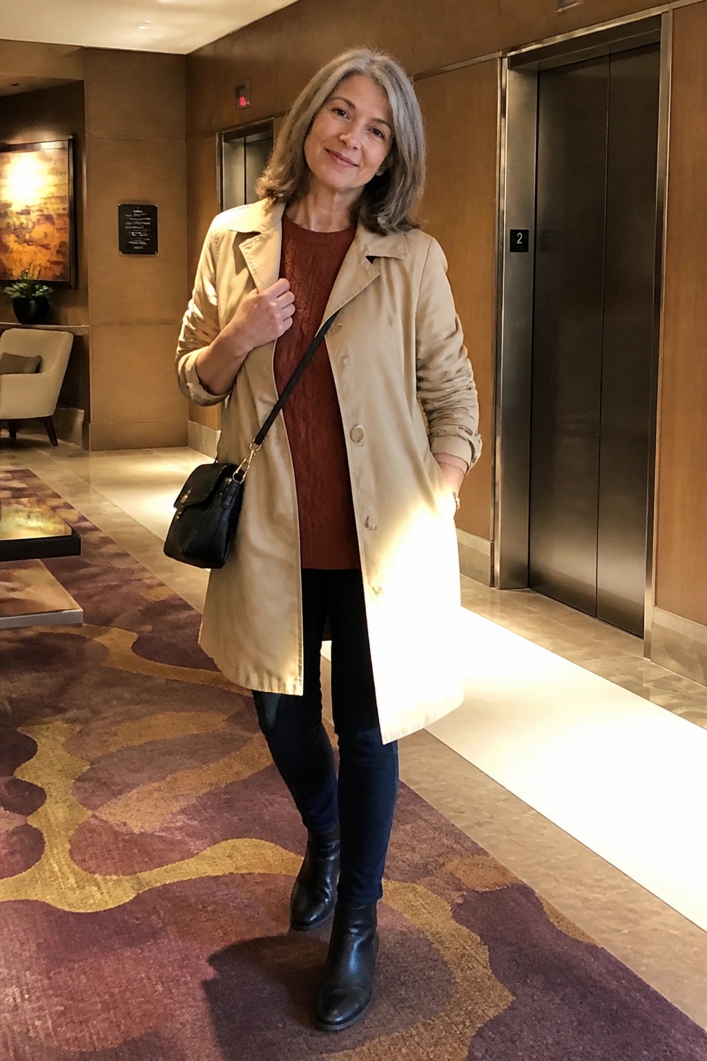 Gray-haired woman in beige trench coat over rust sweater in hotel lobby.