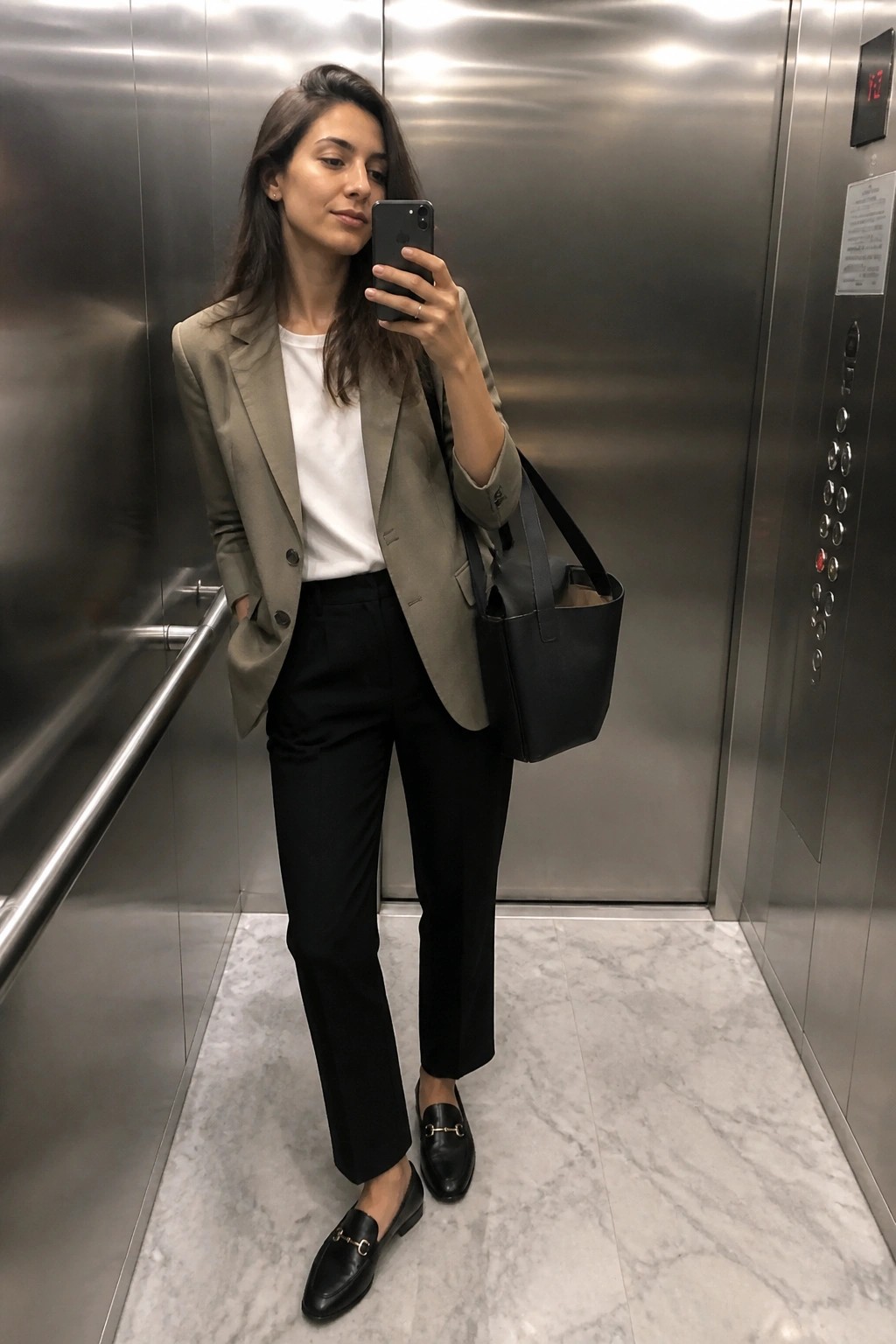 Woman in sage blazer and black pants taking selfie in elevator with tote bag
