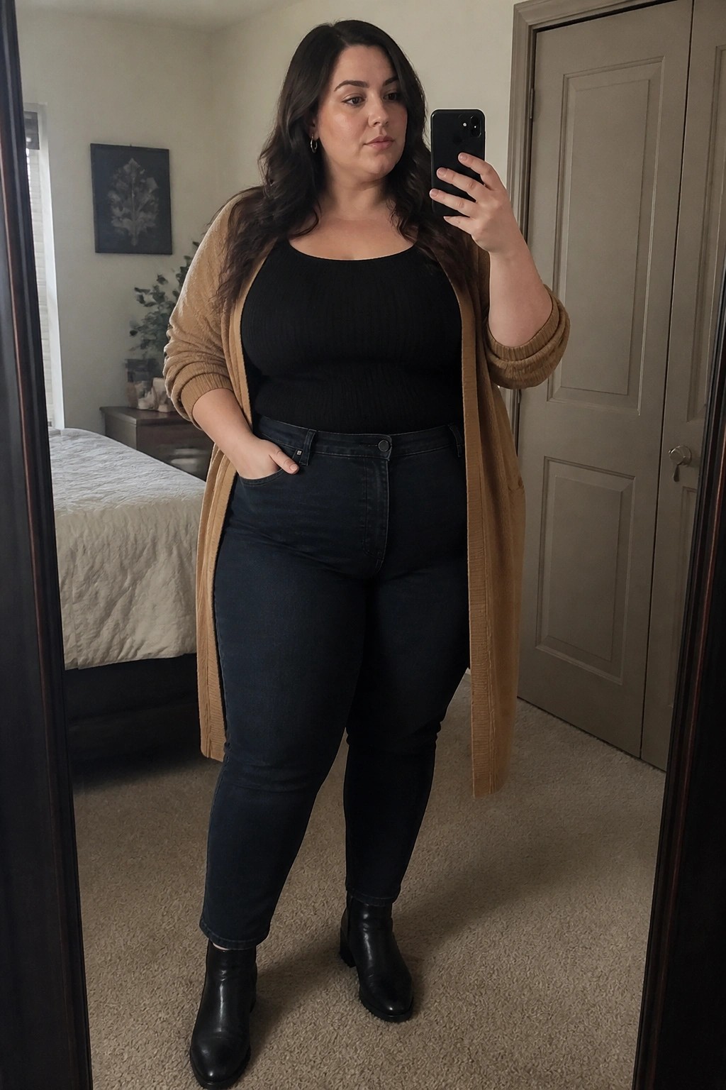 Woman in black top, tan cardigan, and jeans taking mirror selfie in bedroom.