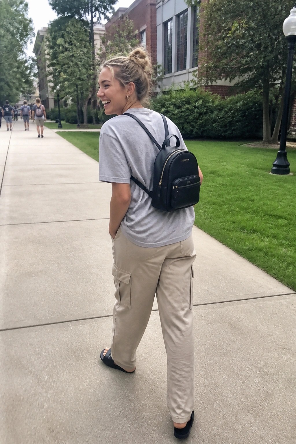 Smiling blonde woman in gray tee and cargo pants with black backpack on campus path
