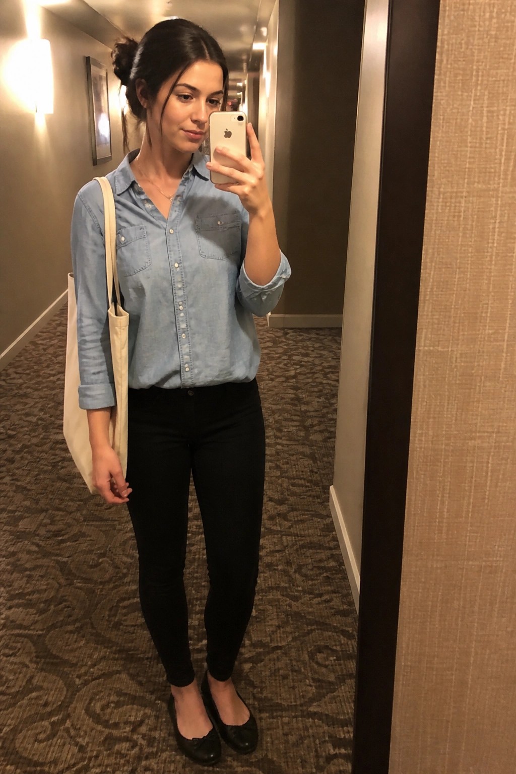 Woman in denim shirt and black jeans takes mirror selfie in hallway with tote bag.