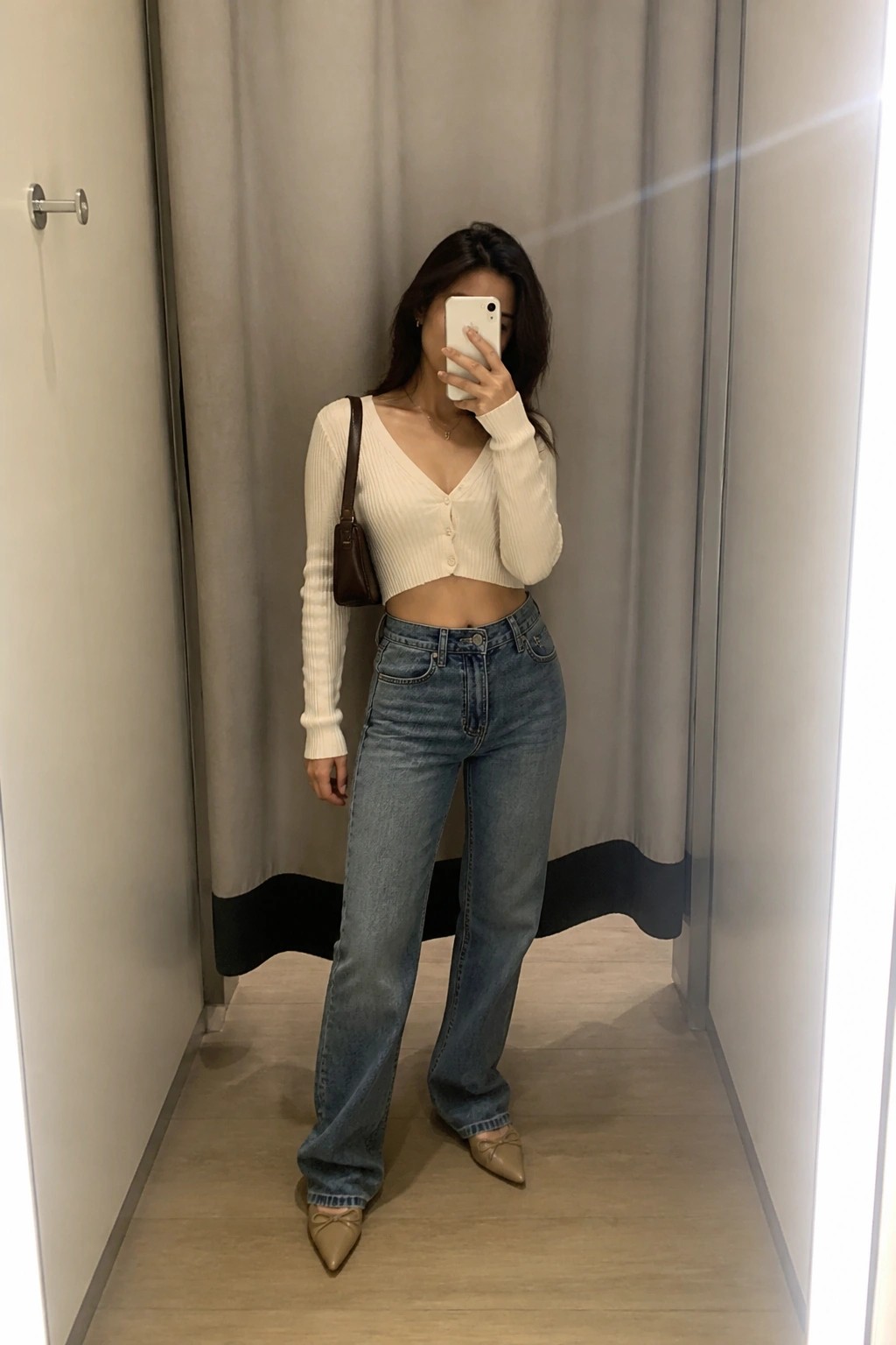 Woman in a white ribbed cropped cardigan sweater with front buttons, blue wide-leg jeans, taupe pointed flats with bows, and a small brown shoulder bag taking a mirror selfie.