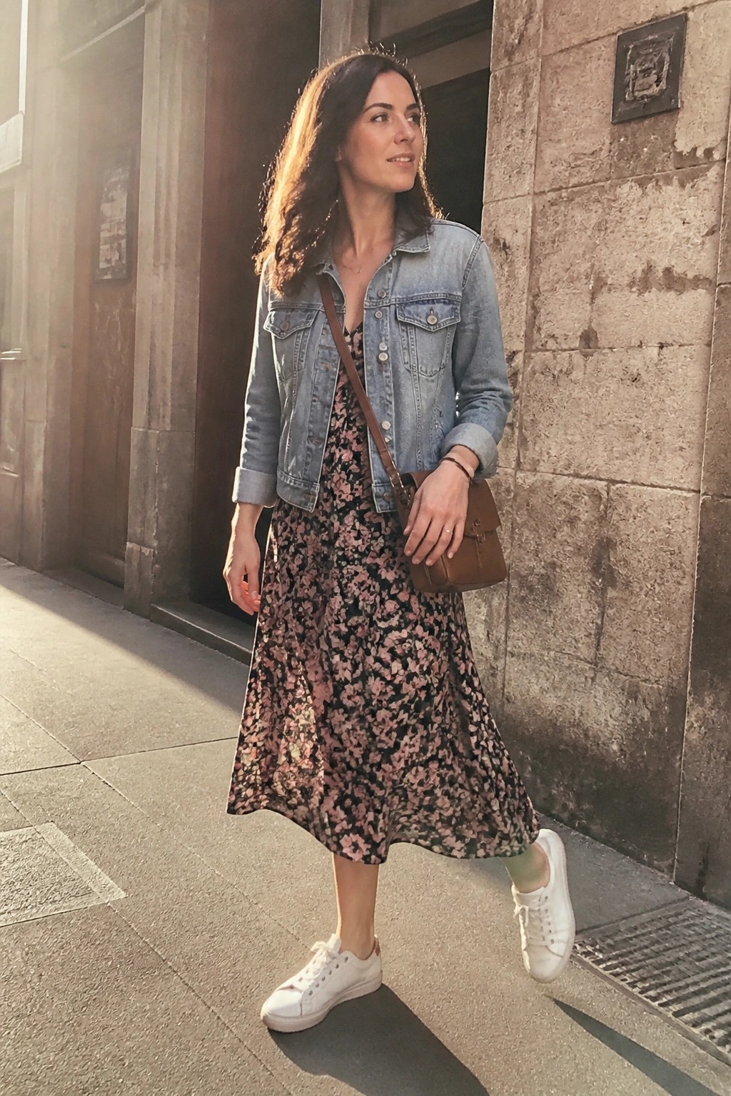 Woman in floral midi dress and denim jacket with white sneakers on sunny street