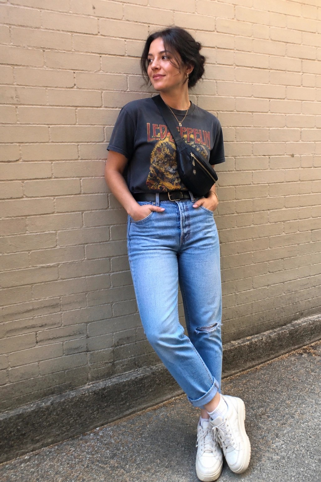 Woman in Led Zeppelin t-shirt, blue jeans and white sneakers by brick wall